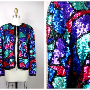 May include: A sequined jacket with a geometric pattern in shades of blue, green, purple, and pink. The jacket has a front zipper closure and long sleeves.