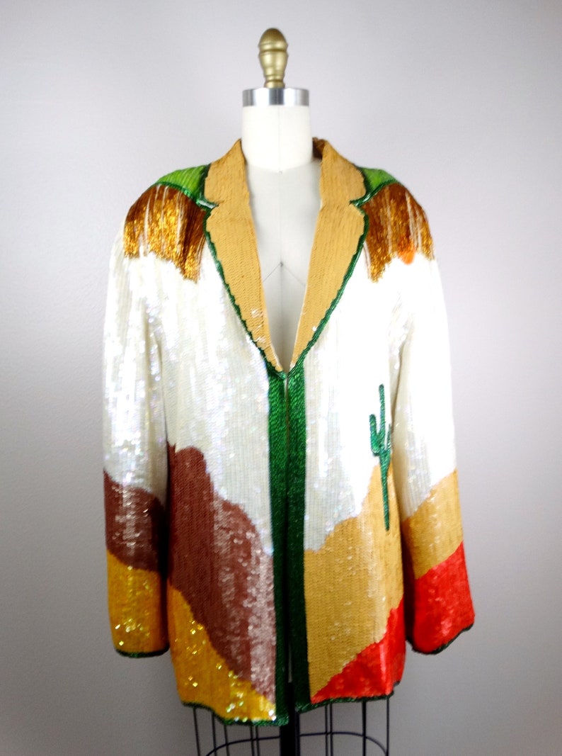 May include: A white sequined jacket with a green, brown, orange, and red desert landscape design. The jacket has a v-neck and a front zipper closure.