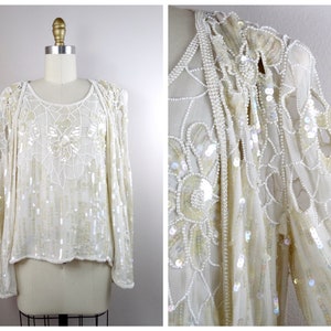 Puede incluir: Una blusa blanca de manga larga con un delicado estampado floral hecho de lentejuelas y cuentas. La blusa tiene un escote redondo y un corte holgado.