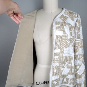 May include: A beige jacket with a white geometric pattern. The jacket is made of a soft, lightweight fabric and has a relaxed fit. The jacket is unbuttoned and the lining is visible.