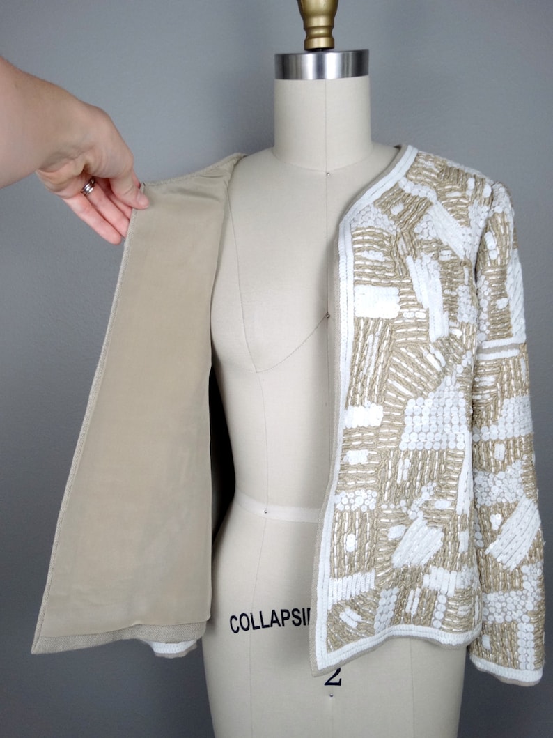 May include: A beige jacket with a white geometric pattern. The jacket is made of a soft, lightweight fabric and has a relaxed fit. The jacket is unbuttoned and the lining is visible.