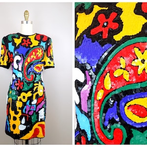 May include: A short-sleeved dress with a black background and a colorful paisley pattern. The dress is covered in sequins and features a variety of colors, including red, yellow, blue, green, and white.