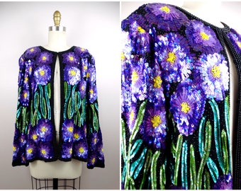 Op de afbeelding: Een vintage jasje versierd met pailletten, met een bloemenontwerp. De jas toont grote paarse bloemen met gele centra en groene stelen tegen een zwarte achtergrond. De randen zijn afgewerkt met zwarte kralen, wat een opvallend visueel contrast creëert.