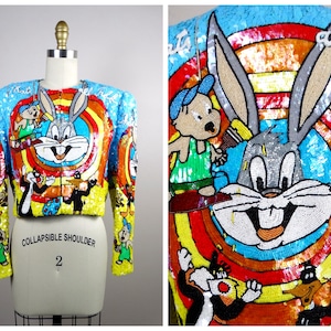 Könnte beinhalten: Eine kurze, mit Pailletten besetzte Jacke mit Bugs Bunny, Tweety Bird, Sylvester der Katze und Porky Pig. Die Jacke ist gelb, blau, rot und grün mit einem Regenbogenmuster. Der Text "COLLAPSIBLE SHOULDER 2" ist auf der Jacke aufgedruckt.