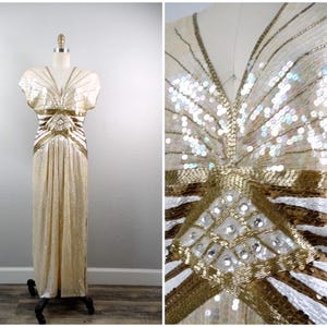 Puede incluir: Un vestido de noche vintage color crema, largo hasta el suelo, sobre un maniquí. El vestido está adornado con lentejuelas y abalorios dorados y plateados. El corpiño presenta un escote en V y mangas cortas. La cintura está resaltada con una banda dorada.
