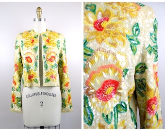 Op de afbeelding: Een crèmekleurige jas met een bloemenpatroon. De jas is gemaakt van pailletten en heeft een inklapbare schouder. De bloemen zijn geel, oranje, rood en groen.