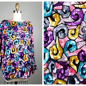 Puede incluir: Una blusa de manga larga con lentejuelas y un estampado abstracto de colores. El estampado presenta remolinos, formas geométricas y una variedad de colores, incluyendo morado, rosa, azul verdoso, dorado y negro.