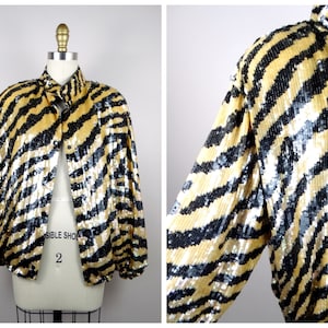 K&ouml;nnte beinhalten: Eine Paillettenjacke mit einem auff&auml;lligen Zebrastreifenmuster in Schwarz und Gold. Die Jacke hat eine Knopfleiste vorne und lange &Auml;rmel. Das Kleidungsst&uuml;ck wird auf einer Schaufensterpuppe pr&auml;sentiert.