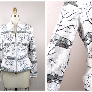 May include: White sequined jacket with a tree branch design. The jacket has a collar, long sleeves, and a silver zipper. Silver sequin bands accent the sleeves and waist. The jacket is displayed on a mannequin.