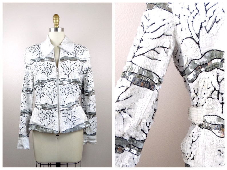 May include: White sequined jacket with a tree branch design. The jacket has a collar, long sleeves, and a silver zipper. Silver sequin bands accent the sleeves and waist. The jacket is displayed on a mannequin.