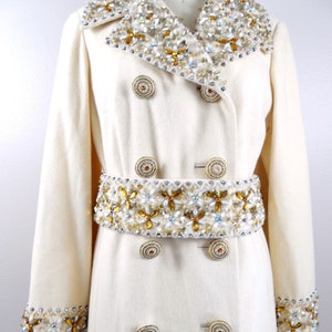May include: A white wool coat with a double breasted closure and a large embellished collar and belt. The embellishments are made of gold, white, and clear beads.