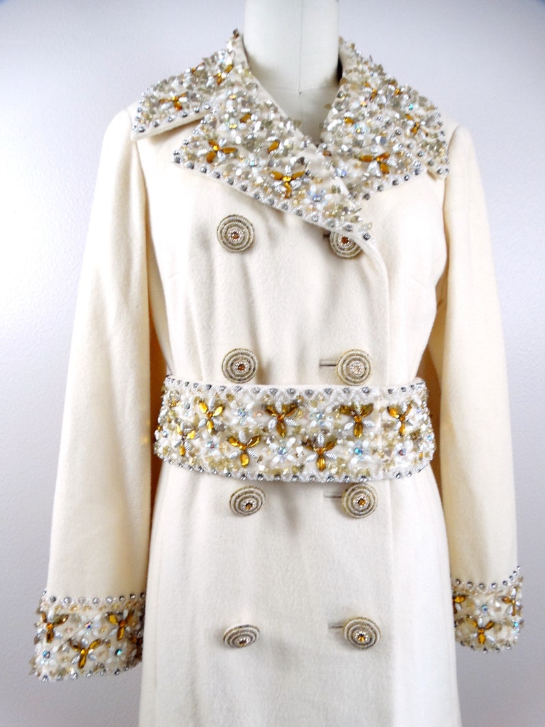 May include: A white wool coat with a double breasted closure and a large embellished collar and belt. The embellishments are made of gold, white, and clear beads.