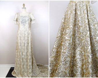 Puede incluir: Un vestido largo blanco con adornos dorados y plateados. El vestido tiene un escote cuadrado y mangas cortas con detalles de flecos. La falda es amplia y fluida.