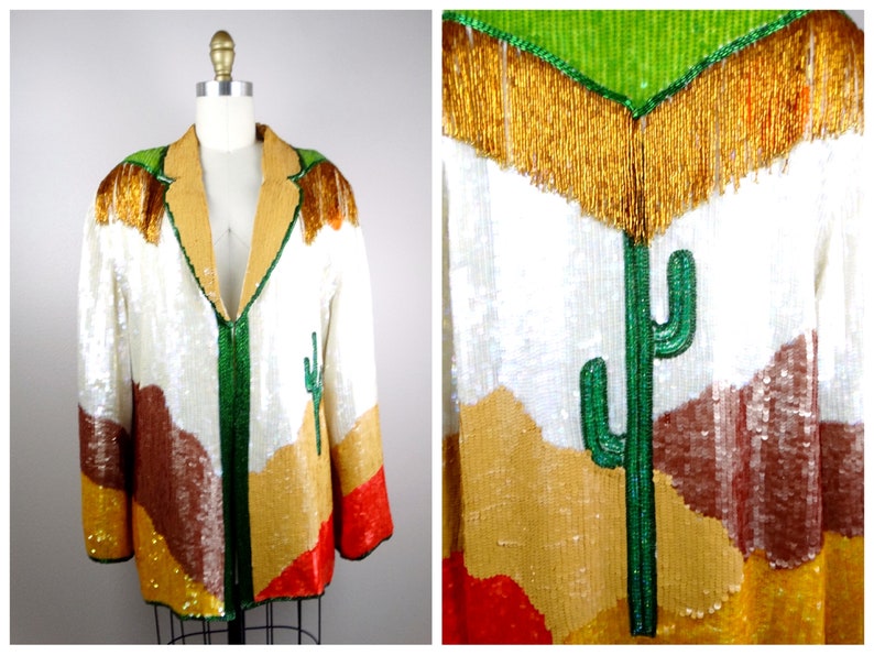 May include: A white sequined jacket with a desert landscape design. The jacket features a green cactus, brown mountains, and gold sequins. The jacket has a green zipper and a gold fringe collar.