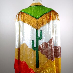 May include: A white sequined jacket with a colorful desert scene design. The scene features a green cactus, brown mountains, and gold sand. The jacket has a green sequined collar with gold fringe.