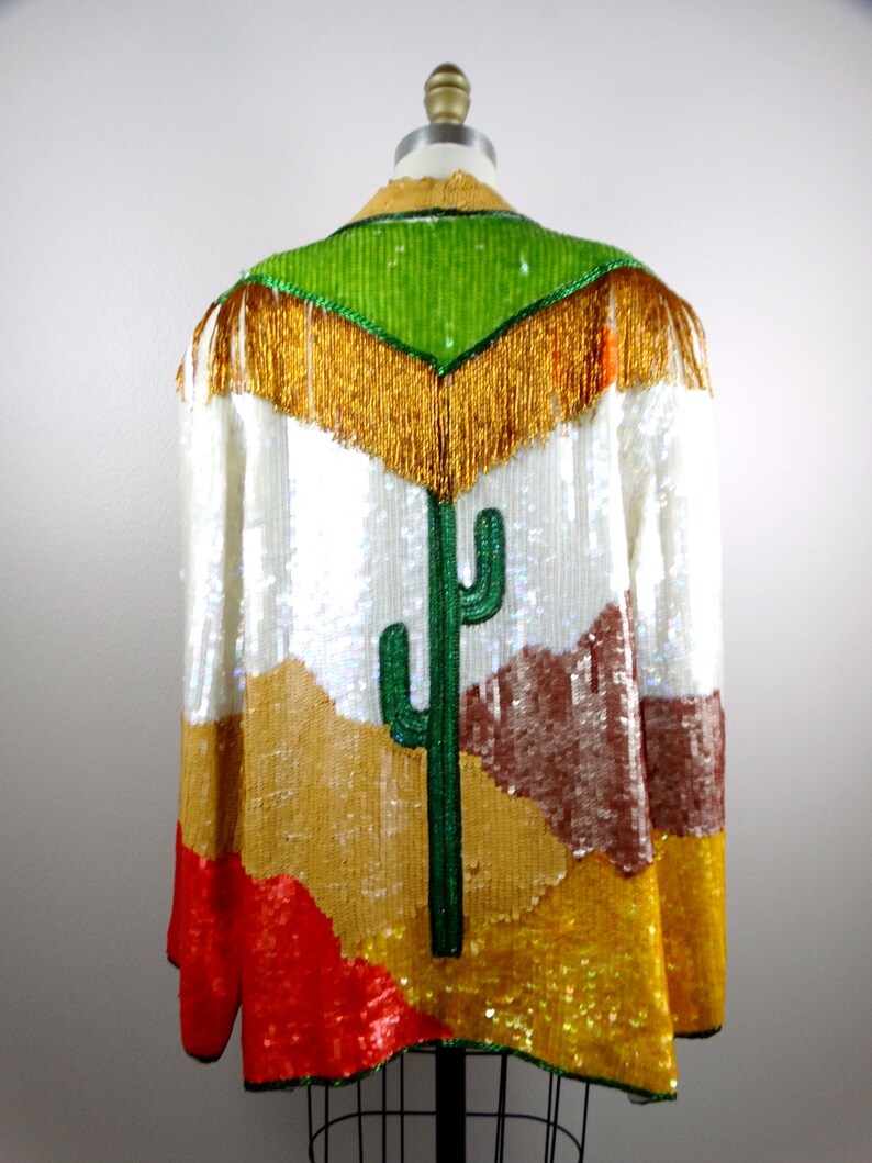 May include: A white sequined jacket with a colorful desert scene design. The scene features a green cactus, brown mountains, and gold sand. The jacket has a green sequined collar with gold fringe.