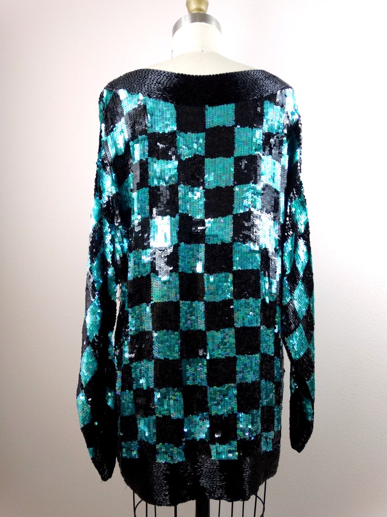 Puede incluir: Una blusa de lentejuelas azul verdoso y negro con un patr&oacute;n de cuadros. La blusa tiene mangas largas y un cuello de barco.