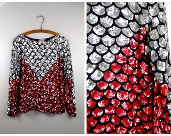 Puede incluir: Una blusa de manga larga con lentejuelas con un patrón de escamas plateadas y rojas. La blusa tiene un escote redondo y mangas largas.