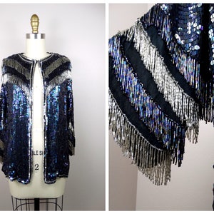Puede incluir: Una chaqueta de lentejuelas con base negra y lentejuelas iridiscentes azules y plateadas. La chaqueta tiene un ribete de flecos alrededor de los bordes y un cuello en V.