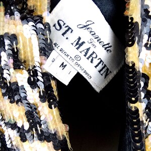 K&ouml;nnte beinhalten: Nahaufnahme eines paillettenbesetzten Kleidungsst&uuml;cks mit einem Etikett mit der Aufschrift "ST. MARTIN" und "Jeanette For". Die Pailletten sind in den Farbt&ouml;nen Schwarz, Gold und schillerndem Wei&szlig; gehalten. Das Kleidungsst&uuml;ck scheint eine Jacke oder ein Oberteil mit schwarzem Futter zu sein.