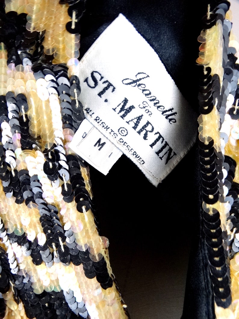 K&ouml;nnte beinhalten: Nahaufnahme eines paillettenbesetzten Kleidungsst&uuml;cks mit einem Etikett mit der Aufschrift "ST. MARTIN" und "Jeanette For". Die Pailletten sind in den Farbt&ouml;nen Schwarz, Gold und schillerndem Wei&szlig; gehalten. Das Kleidungsst&uuml;ck scheint eine Jacke oder ein Oberteil mit schwarzem Futter zu sein.