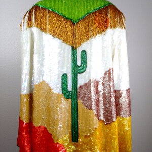 May include: A white sequined jacket with a colorful desert scene design. The jacket features a green cactus, brown mountains, and red and yellow accents. The jacket has a gold fringe trim around the neckline.