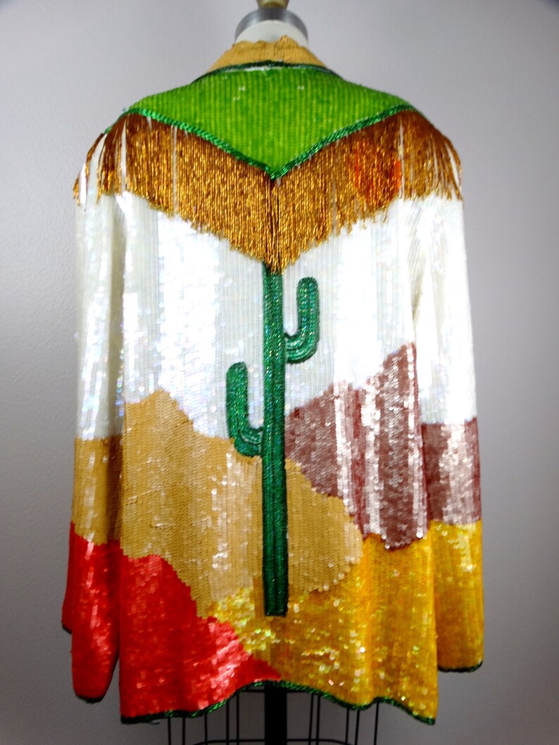 May include: A white sequined jacket with a colorful desert scene design. The jacket features a green cactus, brown mountains, and red and yellow accents. The jacket has a gold fringe trim around the neckline.
