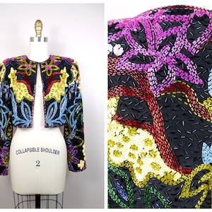May include: A cropped, black sequined jacket with a colorful floral design. The jacket has a collapsible shoulder feature and is labeled "Collapsible Shoulder 2".