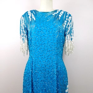 May include: A turquoise blue sequined dress with white beaded fringe on the sleeves. The dress has a round neckline and a fitted silhouette.