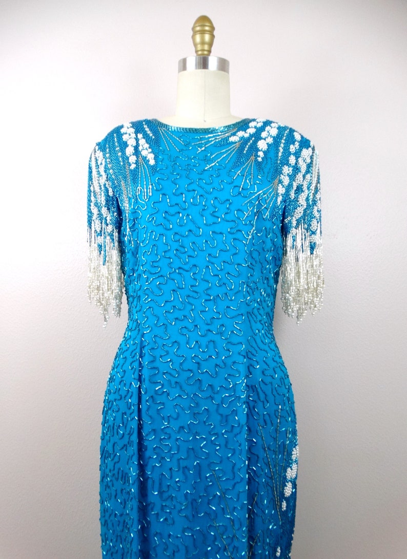 May include: A turquoise blue sequined dress with white beaded fringe on the sleeves. The dress has a round neckline and a fitted silhouette.