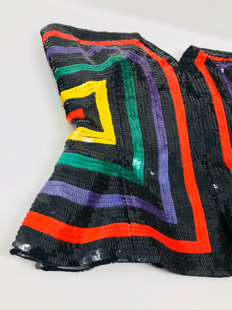 May include: A black fabric with colourful geometric patterns made from sequins. The fabric features red, green, yellow and purple sequins arranged in a geometric design.
