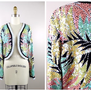 May include: A cropped sequined jacket with a collapsible shoulder. The jacket features a black background with a colorful floral pattern in shades of green, pink, and yellow.
