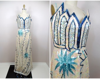 May include: A long, white sequined dress with blue and white floral designs. The dress has a V-neckline and a fitted bodice. The skirt is full and flowing.