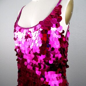 May include: A sleeveless, magenta sequin top. The garment is displayed on a mannequin. The sequins are circular and densely packed, creating a shimmering effect. The straps and neckline are trimmed with a matching magenta fabric.