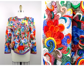 Puede incluir: Una chaqueta colorida y con cuentas con una silueta ajustada y mangas largas. La chaqueta está decorada con bordados intrincados en una variedad de colores, incluyendo rojo, azul, verde, amarillo y blanco.