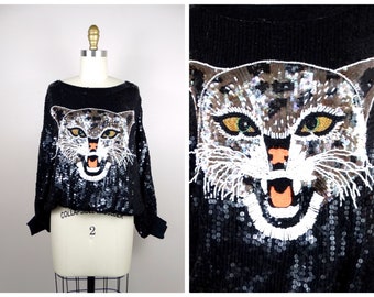 May include: Black sequined sweater with a large embroidered image of a snarling cat's face. The cat's eyes are green and the mouth is open, showing orange teeth.