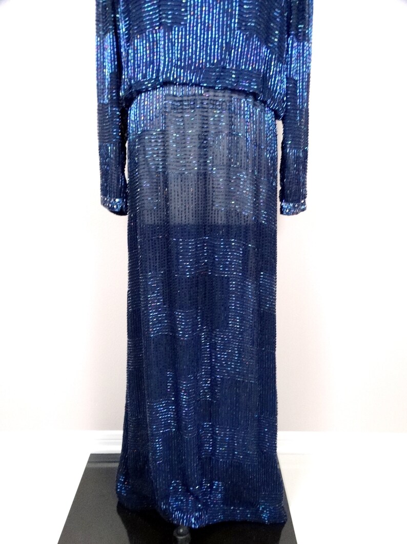 May include: A long, navy blue sequined dress with a fitted bodice and a flowing skirt. The dress is made of small, iridescent sequins that create a shimmering effect.