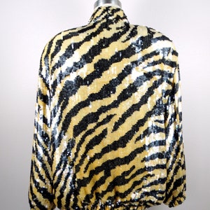K&ouml;nnte beinhalten: Eine Paillettenjacke mit einem Zebrastreifenmuster in Schwarz und Gold. Die Jacke hat lange &Auml;rmel und einen hohen Kragen. Der Saum der Jacke ist mit einem Gummizug versehen. Die Jacke wird auf einer Schaufensterpuppe pr&auml;sentiert.
