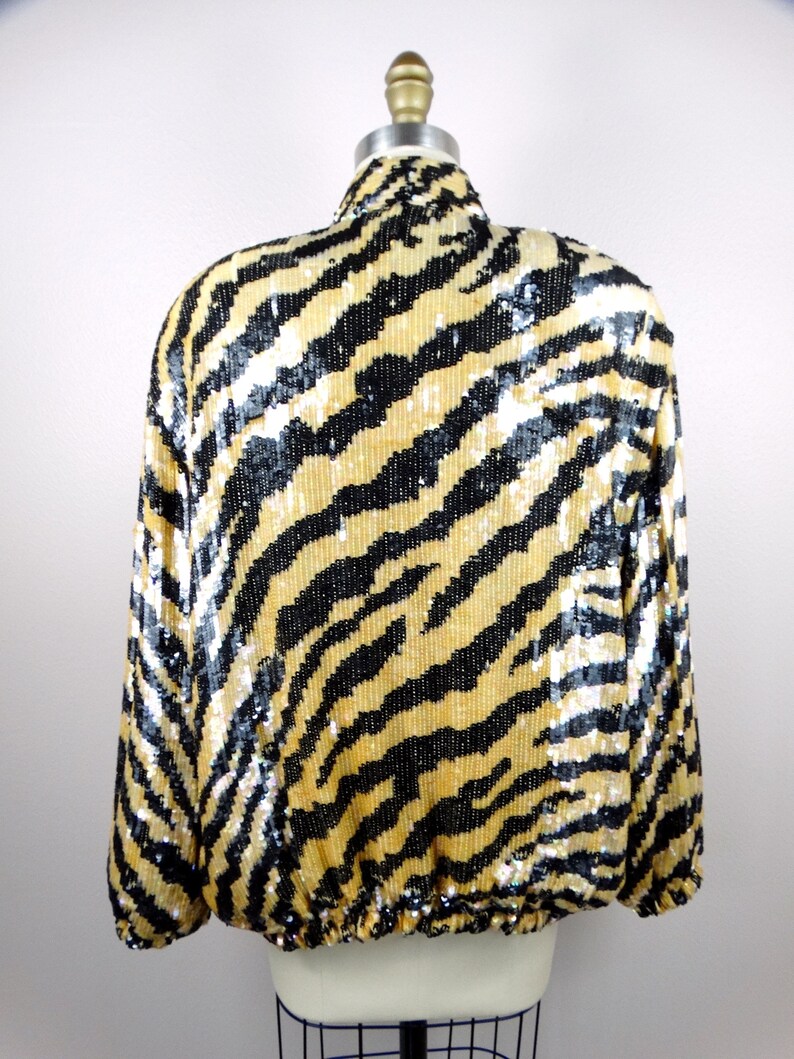 K&ouml;nnte beinhalten: Eine Paillettenjacke mit einem Zebrastreifenmuster in Schwarz und Gold. Die Jacke hat lange &Auml;rmel und einen hohen Kragen. Der Saum der Jacke ist mit einem Gummizug versehen. Die Jacke wird auf einer Schaufensterpuppe pr&auml;sentiert.