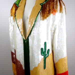 May include: A white sequined jacket with a green, brown, and orange desert landscape design. The jacket has a gold fringe trim and a green cactus embroidered on the front.