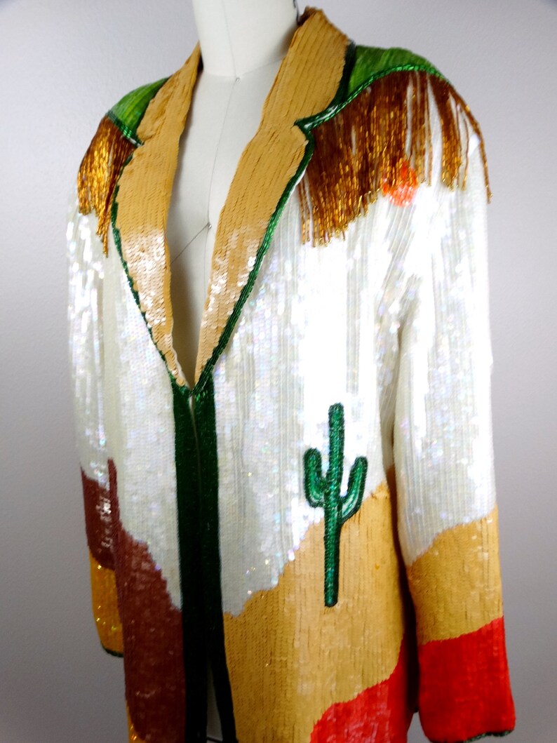 May include: A white sequined jacket with a green, brown, and orange desert landscape design. The jacket has a gold fringe trim and a green cactus embroidered on the front.