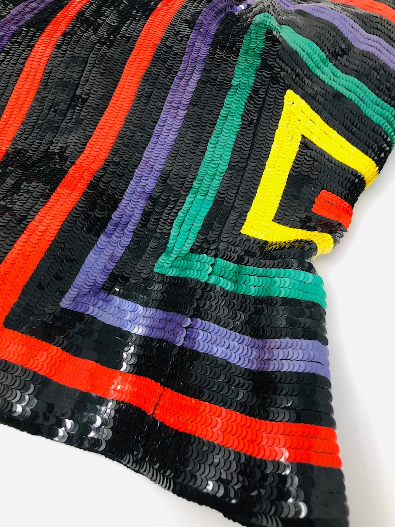 May include: A black fabric with colourful stripes of red, purple, green, and yellow sequins. The sequins are arranged in a geometric pattern.