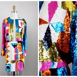 May include: A two-piece sequined outfit, consisting of a jacket and a skirt, displayed on a mannequin. The jacket is open-fronted with long sleeves. The skirt is a mini-skirt. The outfit features a colorful geometric pattern with pink, blue, white, gold, and black sequins.