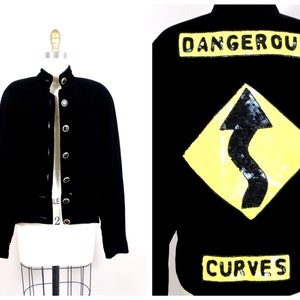 May include: Black velvet jacket with silver button closures and a yellow and black sequined patch that says "Dangerous Curves" with a road sign graphic.
