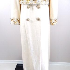 May include: A long, white coat with a double-breasted closure and gold buttons. The coat has a beaded belt with gold and white accents.
