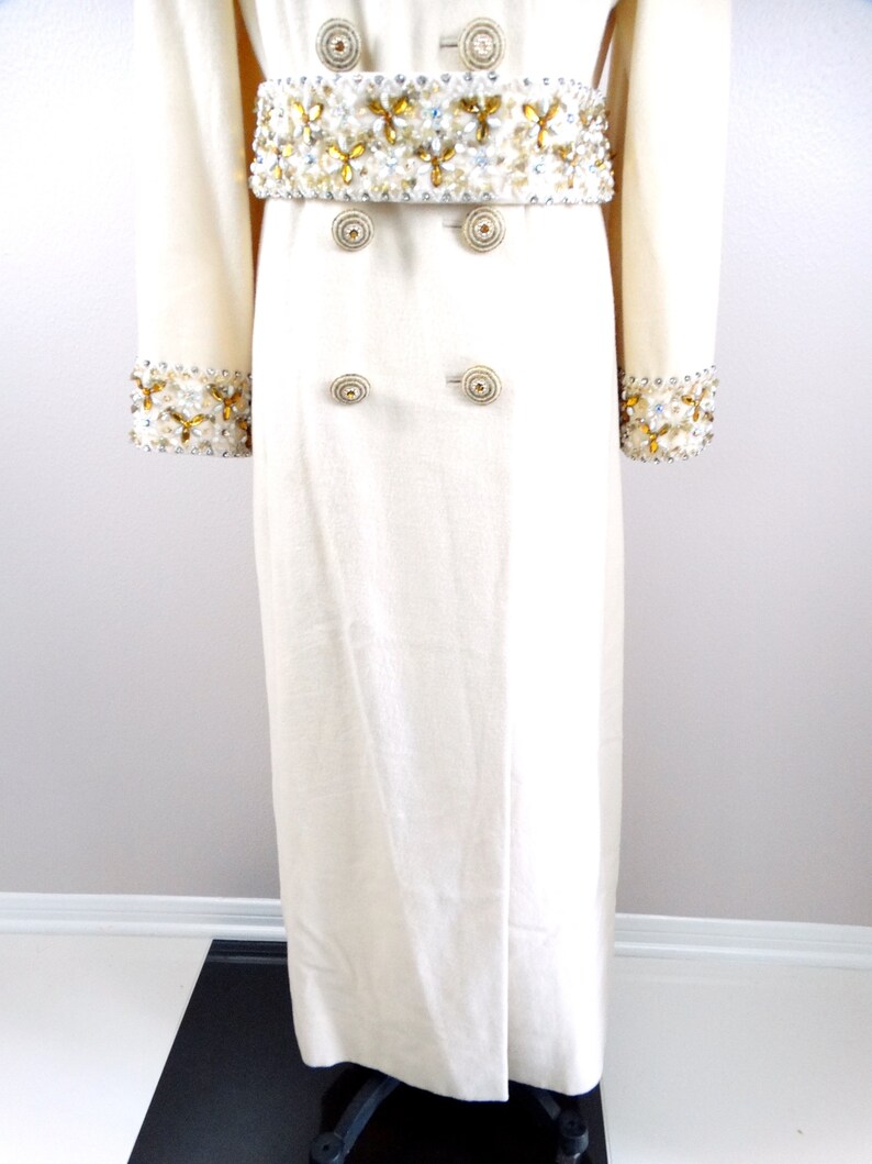 May include: A long, white coat with a double-breasted closure and gold buttons. The coat has a beaded belt with gold and white accents.
