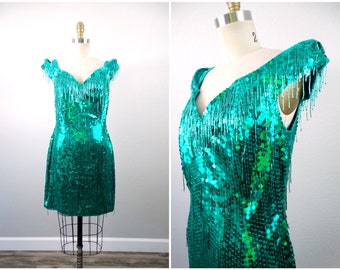 May include: A teal sequined mini dress with fringe detailing on the neckline and sleeves. The dress has a V-neckline and a fitted silhouette.