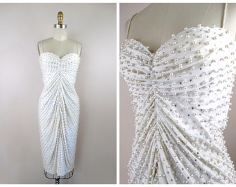 Puede incluir: Un vestido blanco sin tirantes con un corpiño ajustado y una falda fluida. El vestido está cubierto de pequeñas cuentas blancas.