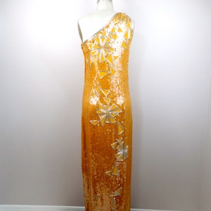K&ouml;nnte beinhalten: Ein langes, einseitig geschnittenes Kleid aus goldenen Pailletten mit silbernen geometrischen Mustern. Das Kleid befindet sich auf einem wei&szlig;en Mannequin.