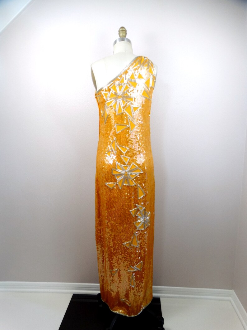 K&ouml;nnte beinhalten: Ein langes, einseitig geschnittenes Kleid aus goldenen Pailletten mit silbernen geometrischen Mustern. Das Kleid befindet sich auf einem wei&szlig;en Mannequin.